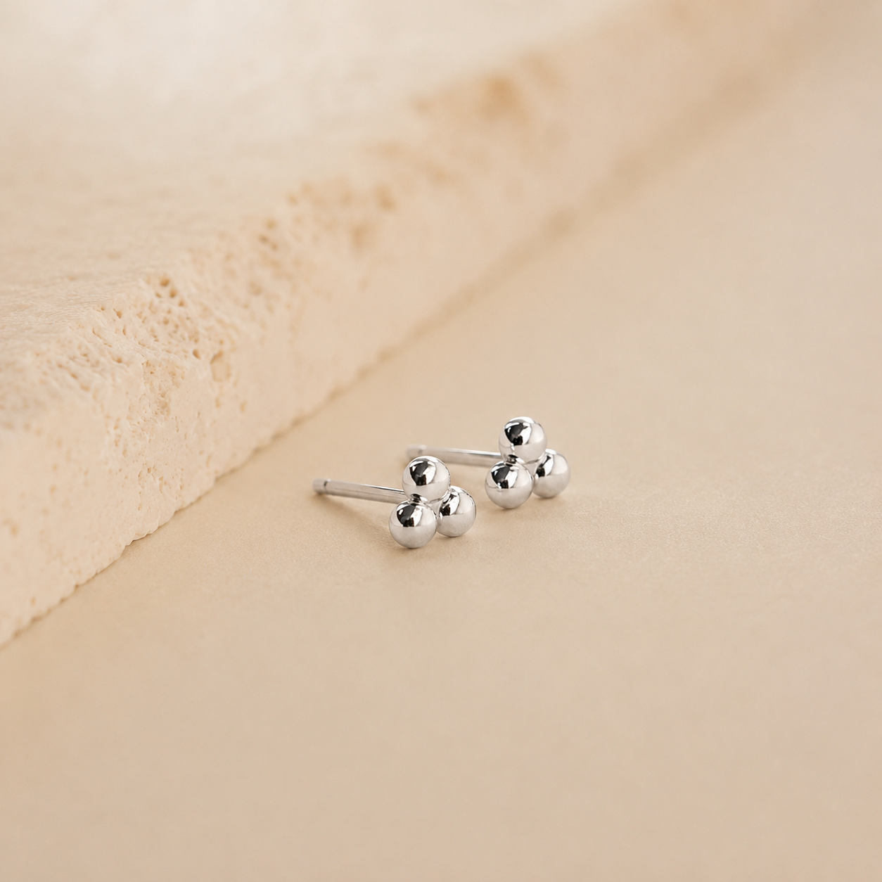 A pair of silver barbell stud earrings with three small polished ball charms on each bar, displayed on a textured beige surface.